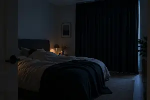 Mørkt soveværelse om natten set fra døren, med en person under en lys dyne set bagfra og et vindue helt dækket af et mørkt gardin